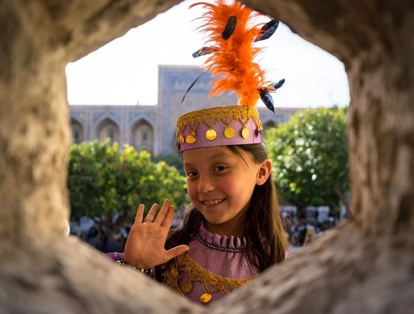 Một bé gái người Uzbekistan trong trang phục truyền thống và giơ tay chào khi du khách giơ máy ảnh lên chụp.