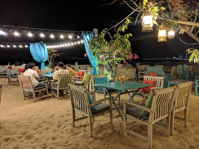 19h30: Nhâm nhi đồ uống ở Surf Bar  Nghỉ ngơi sau một ngày khám phá thành phố biển, Surf Bar là điểm đến lý tưởng tiếp theo với điểm cộng là những bộ bàn ghế gỗ đặt ngay trên nền cát, nằm trên đường ven bờ biển Xuân Diệu. Ngồi thưởng thức những món đồ uống tại quán, du khách có thể tận hưởng không gian thoáng đãng với những làn gió biển mát rượi, tiếng sóng vỗ rì rào. Một số đồ uống như cocktail, nước ép đều có giá dưới 40.000 đồng. Ảnh: Vy An.